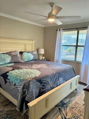 Ein Schlafzimmer mit Bett, Deckenventilator, Nachttisch, Lampe und einem Fenster mit Vorhängen.