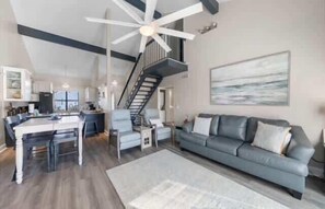A modern living room with a gray sofa, a dining area with a white table, and a staircase leading to an upper level.