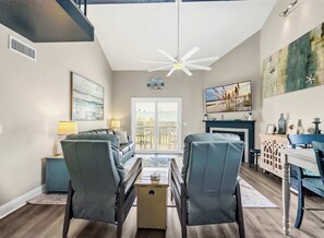 A modern living room with a ceiling fan, a fireplace, and a large window. - Sandy Toes Retreat @ Mariner Lakes – Beautiful Condo with Easy Beach Access! (Orange Beach)