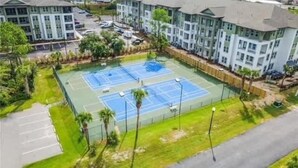 Una cancha de tenis rodeada por una valla, con un estacionamiento y edificios al fondo.