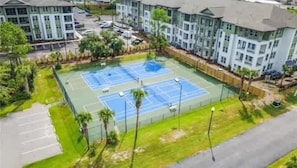 Ein Tennisplatz, umzäunt, mit einem Parkplatz und Gebäuden im Hintergrund.