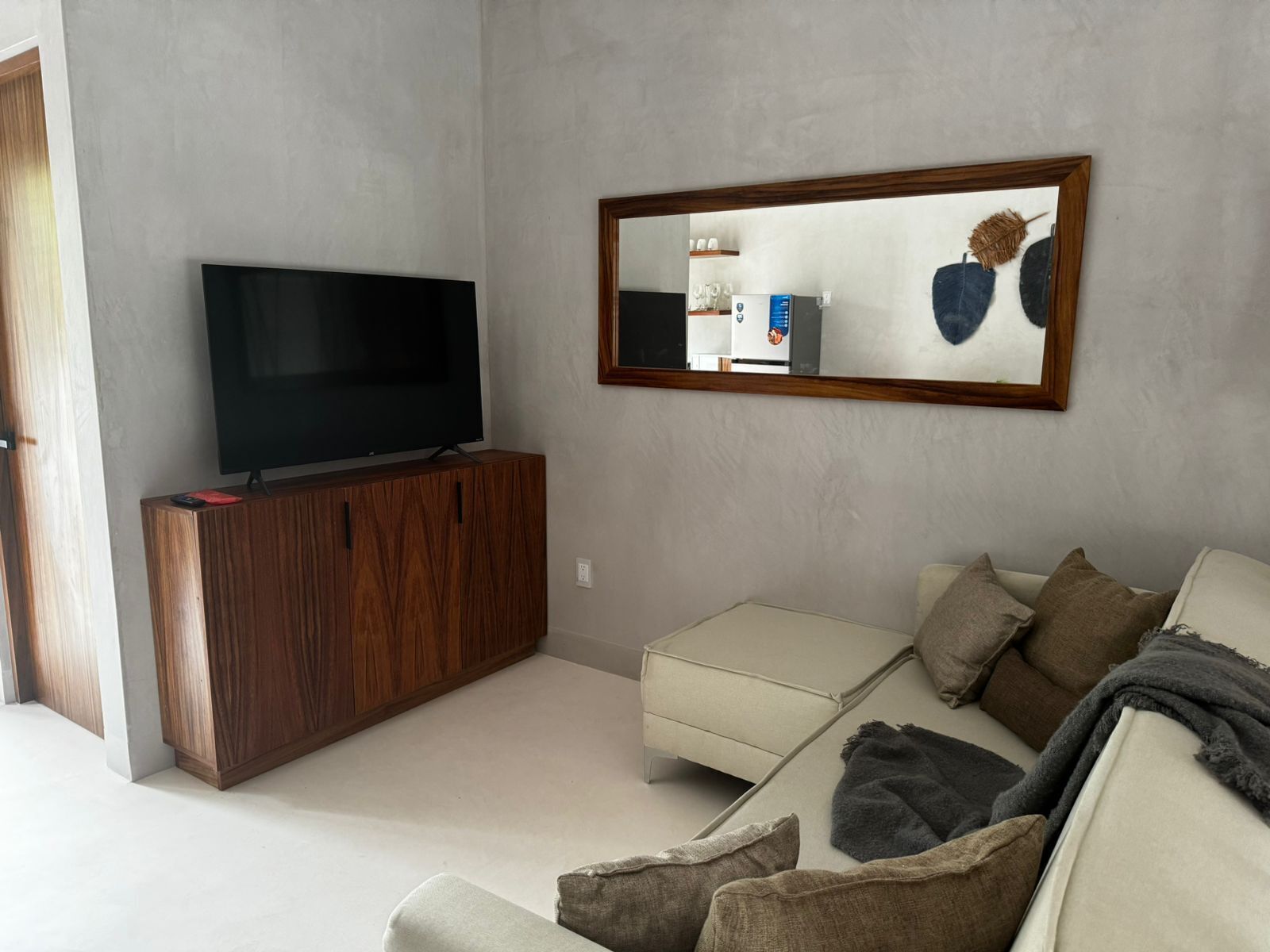 A modern living room with a flat-screen TV on a wooden cabinet, a cream-colored sofa, and a large mirror on the wall.