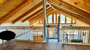 Une chambre spacieuse située dans les combles, avec des poutres en bois, une chaise noire, deux lits, un escalier et une vue sur l’extérieur.