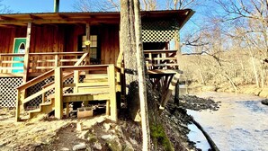 Un chalet rustique avec une façade en bois, une cheminée, une véranda dotée d’une balustrade en treillis et une vue sur une rivière.