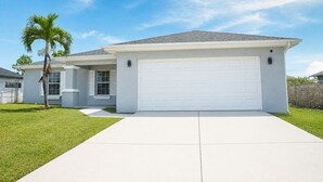 Een gelijkvloers huis met een witte garage, een kleine voortuin met een palmboom en een helderblauwe lucht.