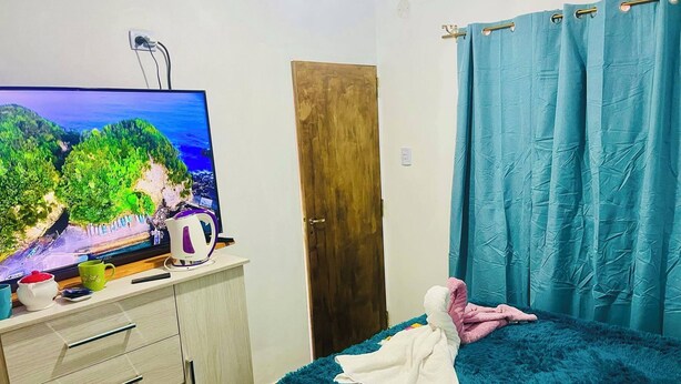 Une chambre à coucher avec un lit, un téléviseur posé sur une commode en bois, une porte et un rideau.