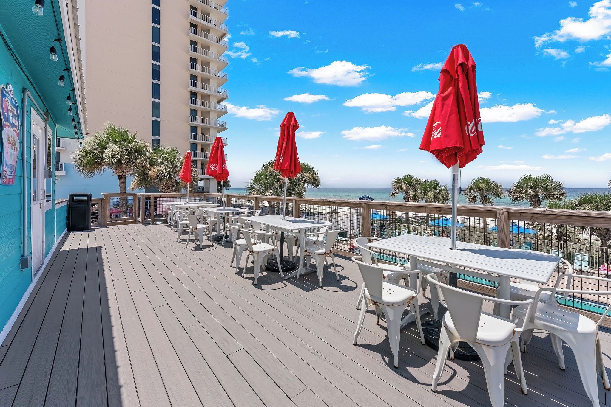 A deck with outdoor seating and red umbrellas, overlooking a pool and palm trees.