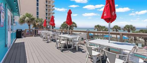 A deck with outdoor seating and red umbrellas, overlooking a pool and palm trees.