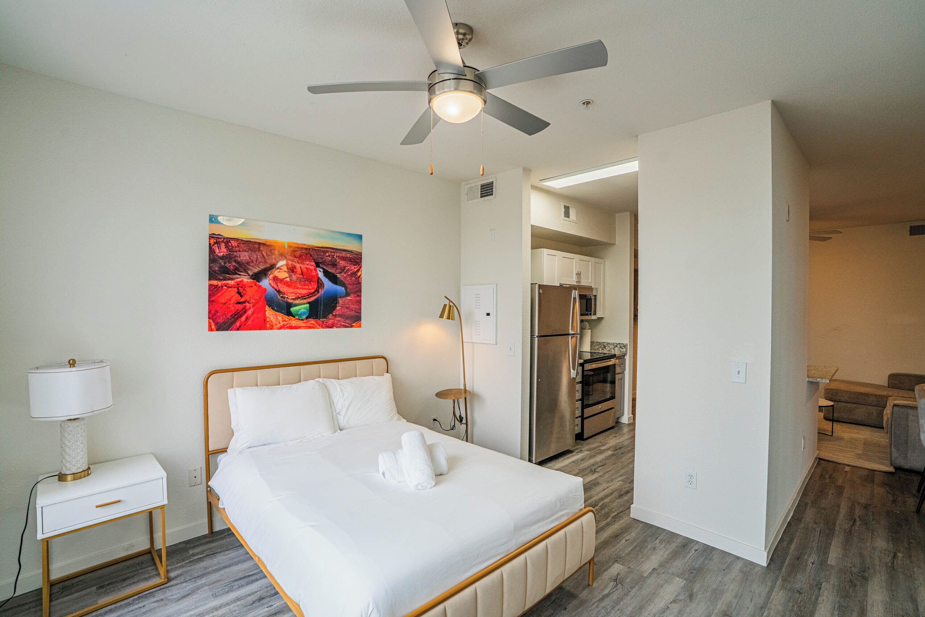 A hotel room with a bed, a ceiling fan, a painting, a nightstand, and a view into the kitchen area.