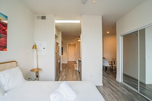 Quarto de hotel com cama, escrivaninha, cadeira, abajur e vista para uma cozinha e área de refeições.