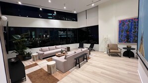 Ein modernes Wohnzimmer mit einer großen Couch, einem Couchtisch und einem Beistelltisch. Eine Topfpflanze und eine Lampe sind ebenfalls vorhanden. Das Zimmer verfügt über große Fenster mit Blick auf die beleuchtete Stadt bei Nacht.