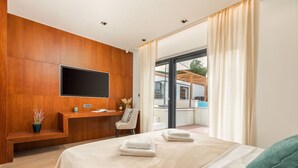 A modern hotel bedroom with a large bed, a flat-screen TV mounted on a wooden panel wall, a small desk with a chair, and a view of the outdoors through large windows. - Luxurious Villa Istria Green with private pool - for families and nature lovers (Boljevići)