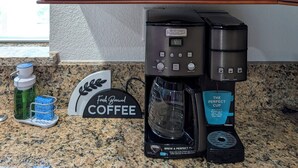 Una cocina con cafetera, un letrero que dice 'Café recién hecho' y una repisa con vajilla y tazas blancas.