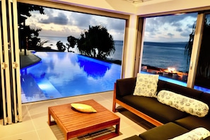 Un moderno salón con un amplio ventanal corredera que da a una piscina infinita privada con vistas al océano.