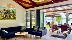 A modern living room with a dark blue sofa, a white coffee table, and a ceiling fan. There is a large window with metal shutters, and a view of the beach is visible outside.