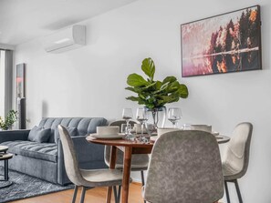 Un salon moderne avec un canapé gris, une table à manger avec des chaises, une grande plante et un tableau représentant une forêt accroché au mur.