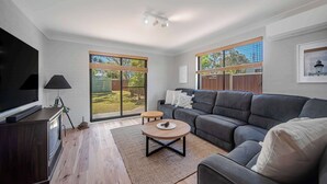 Ein modernes Wohnzimmer mit einem Flachbildfernseher, einem grauen Sofa, einem runden Couchtisch und einer großen Glasschiebetür, die zu einem Garten führt.