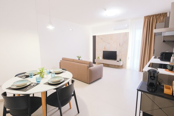 A modern hotel room with a dining area, a sofa, a TV, and a kitchen area.