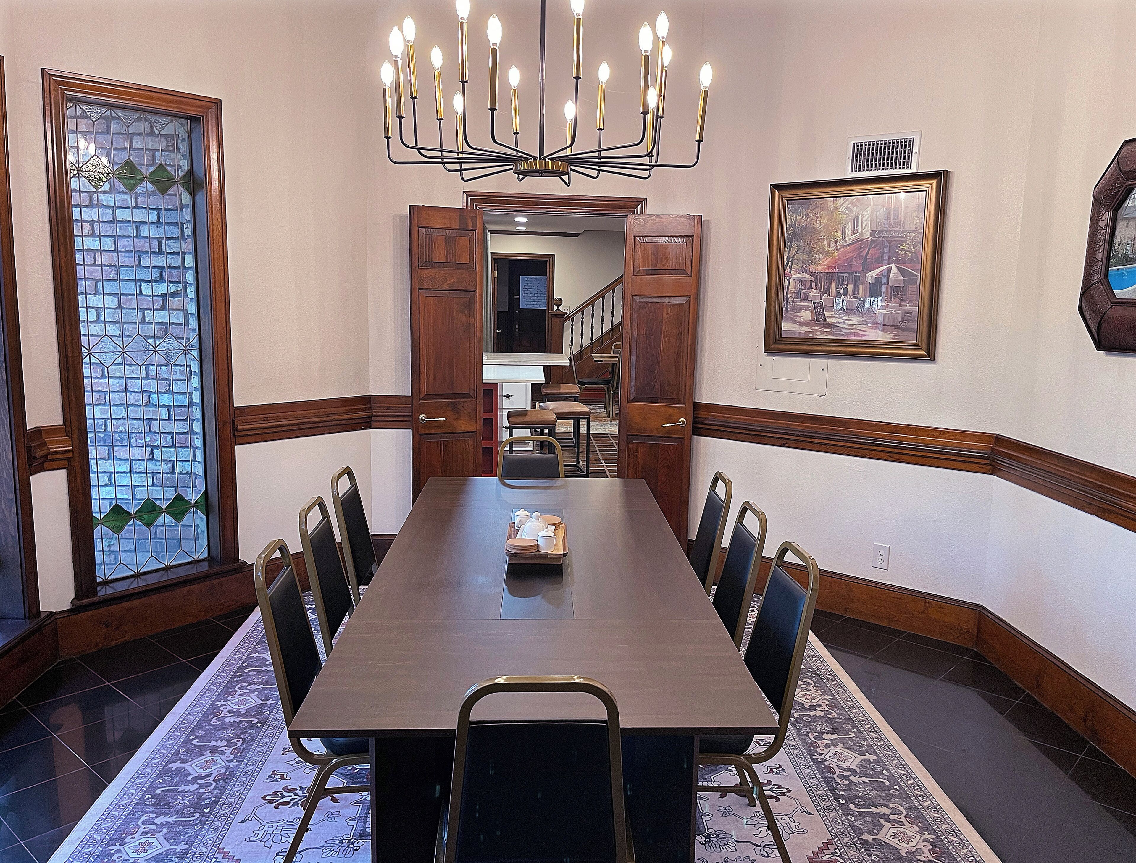 Uma sala de conferências com uma longa mesa, cadeiras, um candelabro, uma janela de vitrais e um quadro na parede.