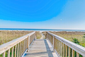 Ein hölzerner Steg führt zu einem Strand unter blauem Himmel.