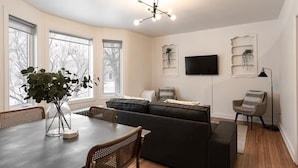 A modern living room with a dining table, black leather sofa, and a TV mounted on the wall. - Home-Style 1BR Near HSC | Free Parking (Winnipeg)