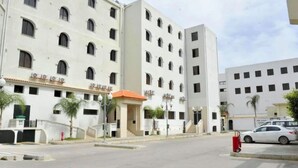 A multi-story hotel with a central entrance, multiple balconies, and a parking area with a white car. - Le Numidien - Aeroport (Algiers)