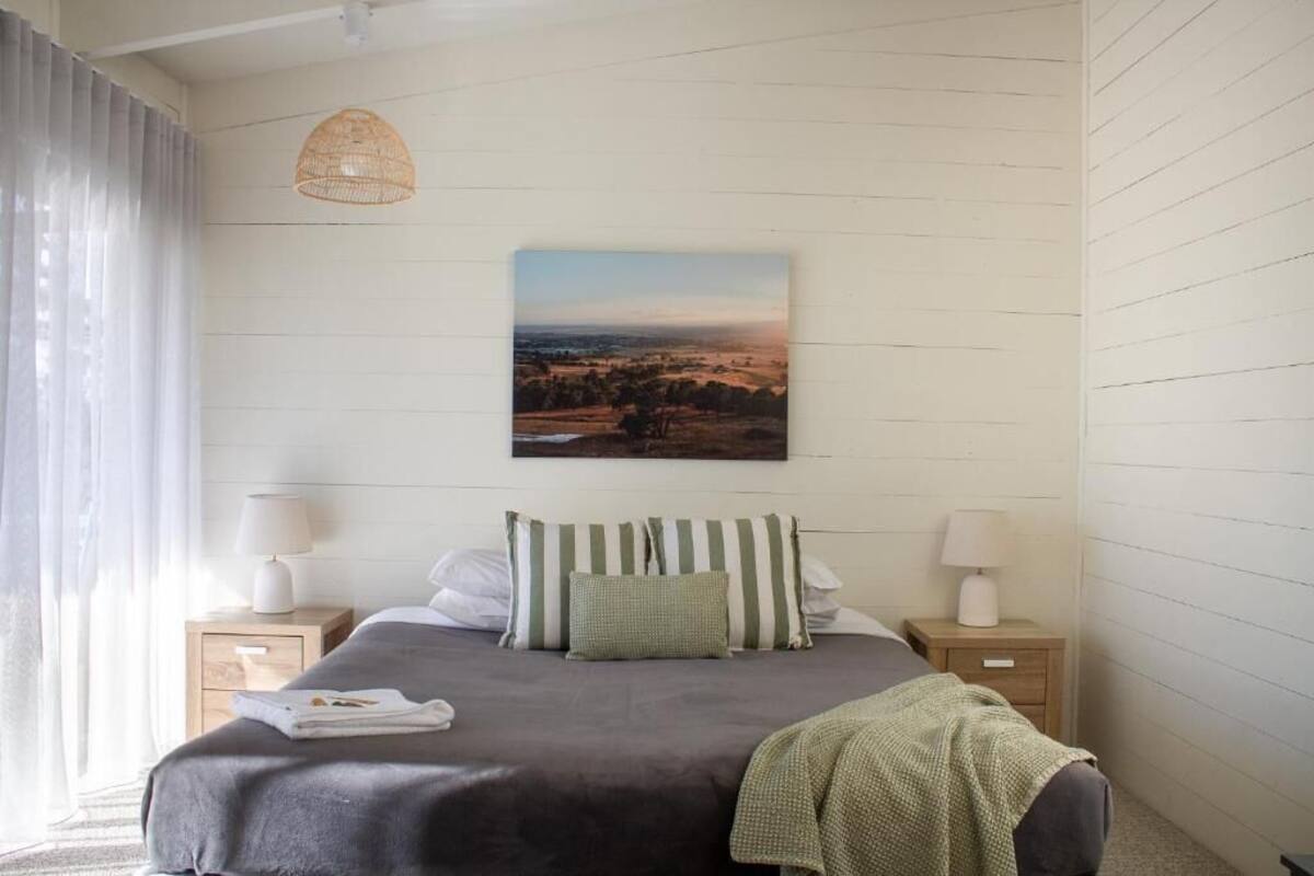 A bedroom with a bed, two bedside tables, a hanging lamp, and a landscape painting on the wall.