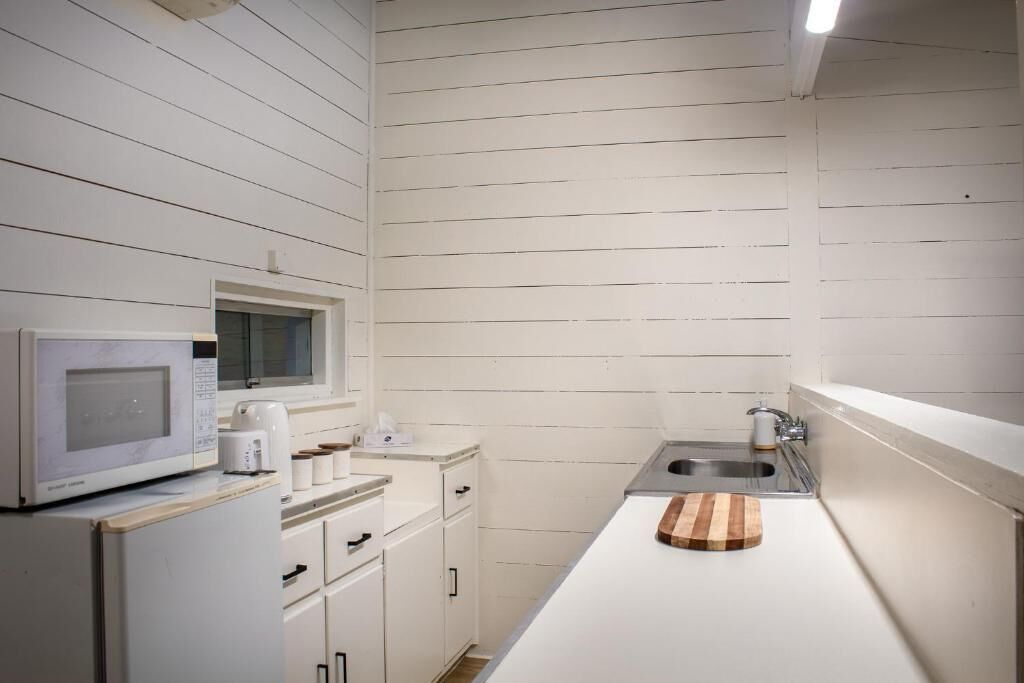 Een kleine keuken met witte kastjes, een magnetron, een spoelbak en een houten snijplank.