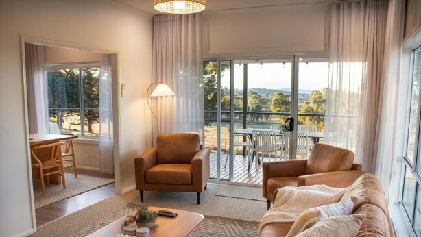 En moderne stue med sofa, lænestol og sofabord. Der er en balkon med bord og stole, samt udsigt til naturen.