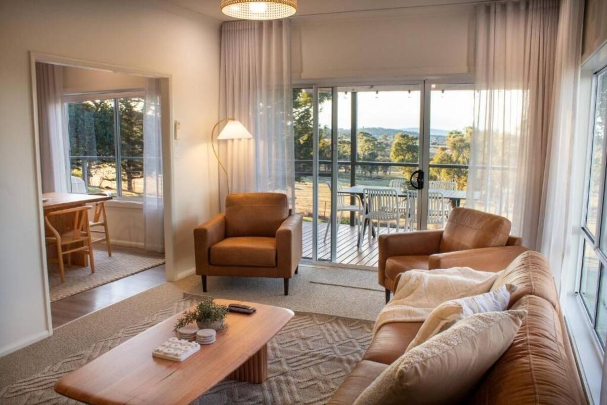 A modern living room with a sofa, armchair, and coffee table. There is a balcony with a table and chairs, and a view of the outdoors.