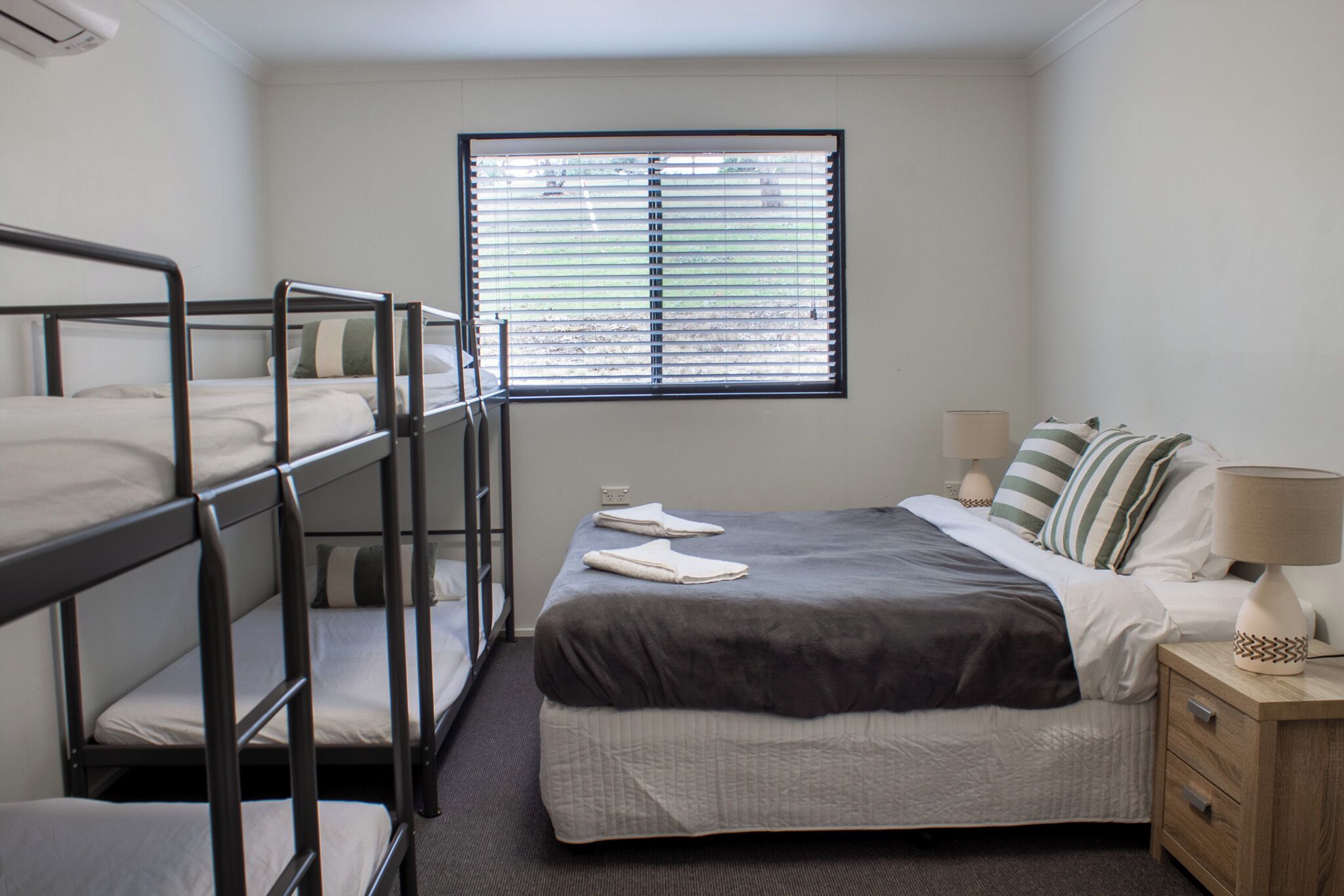 Una habitación con literas, una cama individual, una ventana con persianas, una lámpara de noche y una mesita de noche.