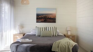 A bedroom with a bed, two bedside tables, a hanging light, a painting, and a window with curtains.