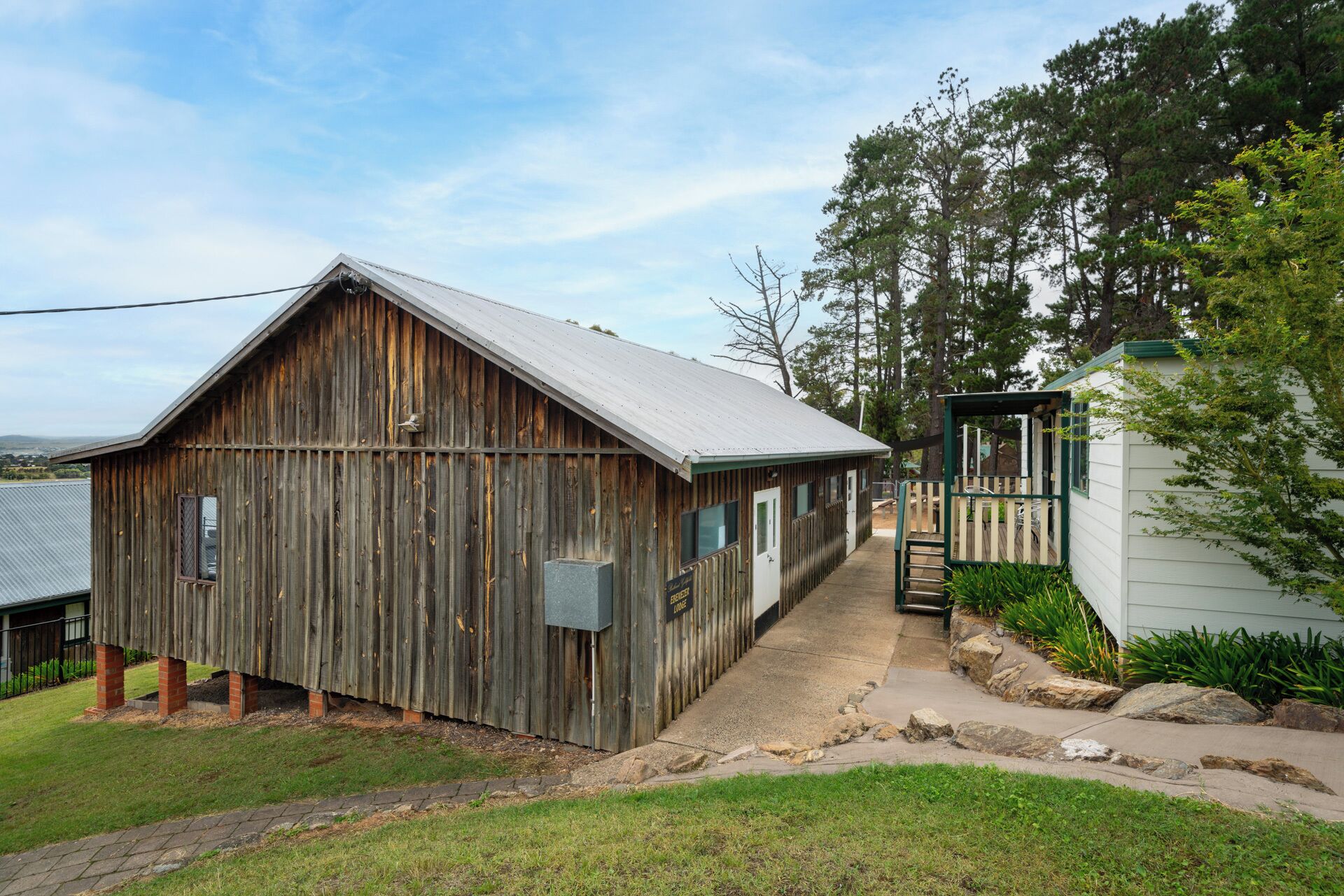 A rustic wooden barn with a metal roof, a small adjacent structure with a green door, and a paved pathway leading to a gated area.