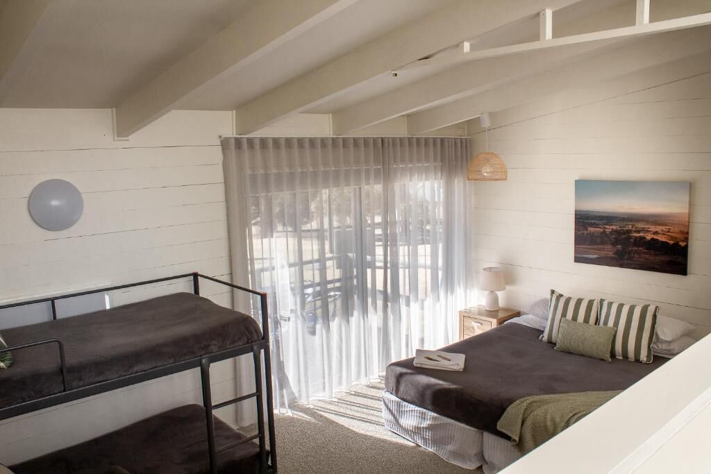 Une chambre avec un lit superposé, un lit simple, une table de chevet avec une lampe, une fenêtre avec des rideaux légers et un tableau accroché au mur.