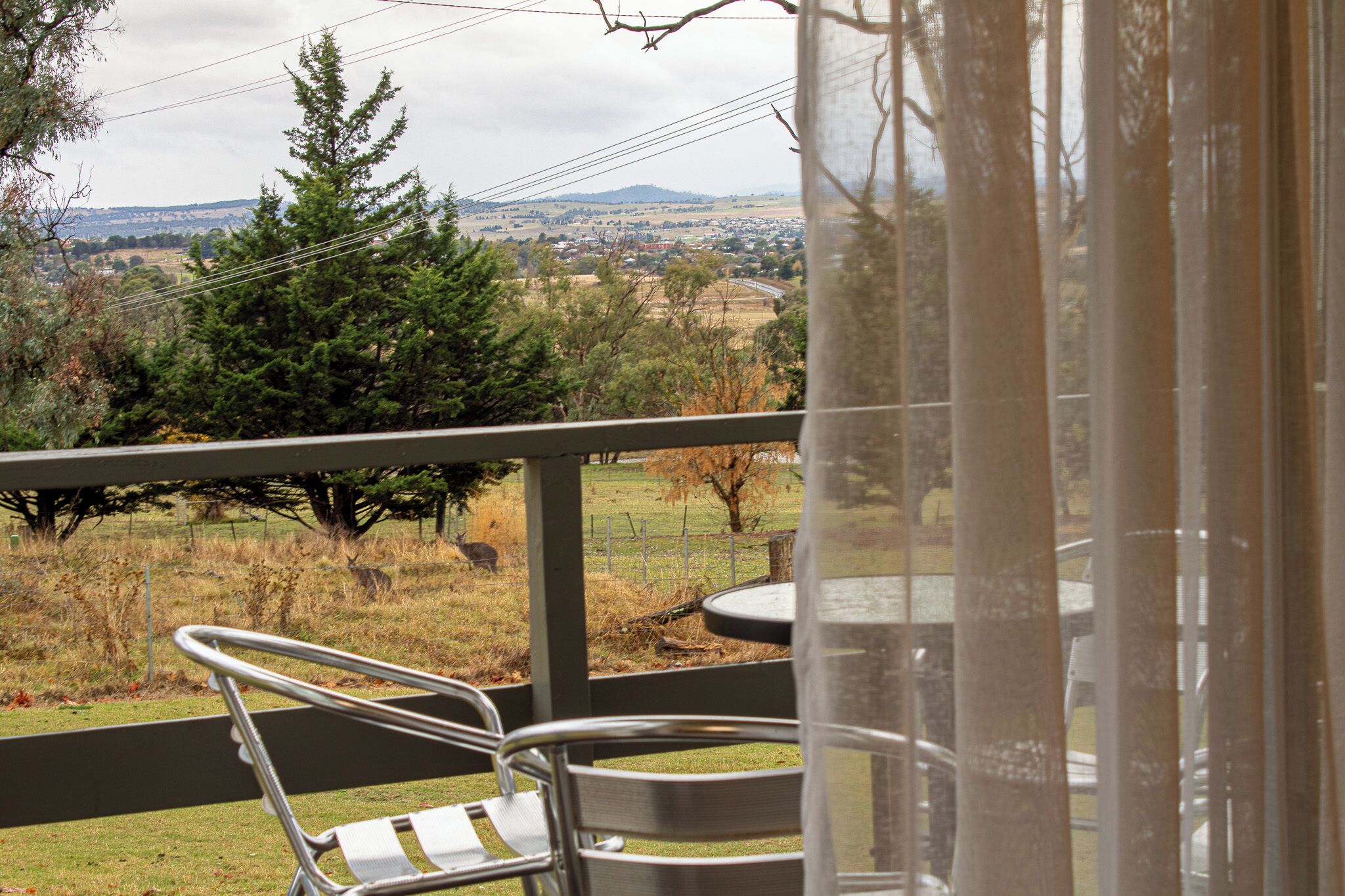 A balcony with a metal chair and table, overlooking a grassy area with trees and a distant town.