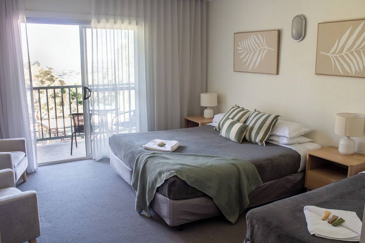 A hotel room with a balcony, a bed with a green blanket, two bedside tables with lamps, and two framed leaf designs on the wall.