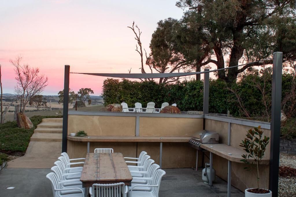 An outdoor patio with a wooden table and white chairs, a barbecue grill, and a view of the landscape.
