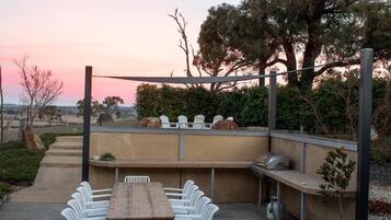 An outdoor patio with a wooden table and white chairs, a barbecue grill, and a view of the landscape.