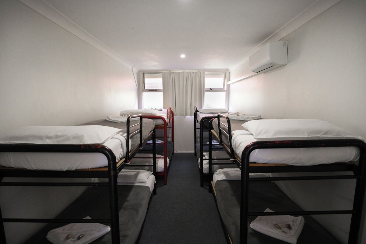 A room with four bunk beds arranged in a row, a central window with curtains, and an air conditioning unit on the wall.