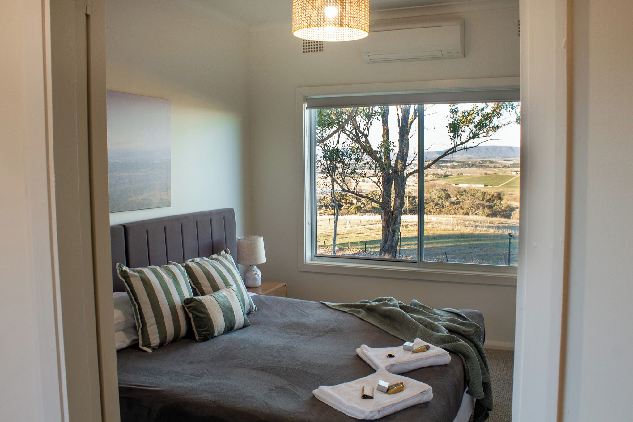 A bedroom with a large window, a bed with striped pillows, and a view of a tree and landscape.