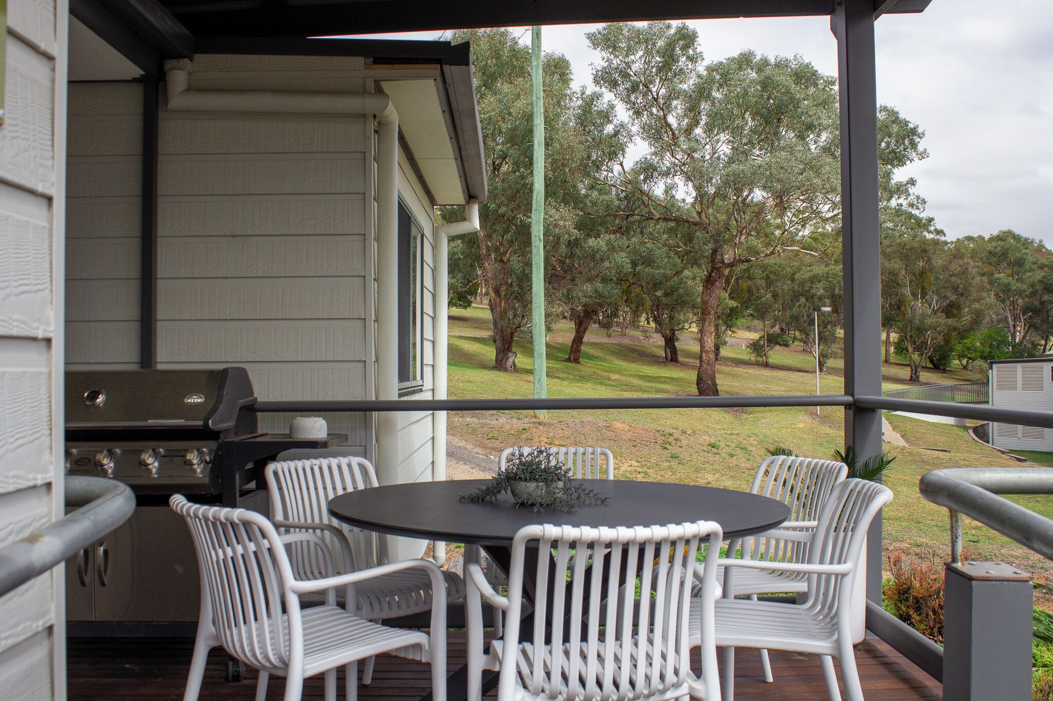 An outdoor patio with a grill, white wicker chairs, and a view of a grassy area with trees.