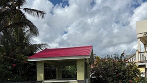 A covered outdoor seating area with a red roof, surrounded by lush greenery and a paved walkway.