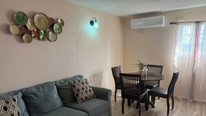 A living room with a sofa, patterned cushions, a dining table with chairs, a wall-mounted air conditioner, and a window with curtains.