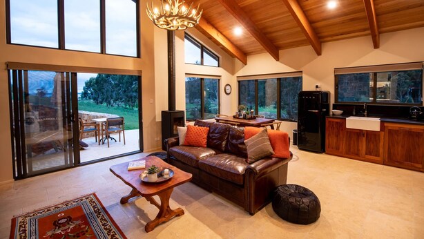 A spacious living room with a leather sofa, wooden coffee table, and a chandelier. Large windows offer a view of the outdoors. - Bruny Island Haven - The Studio (Adventure Bay)