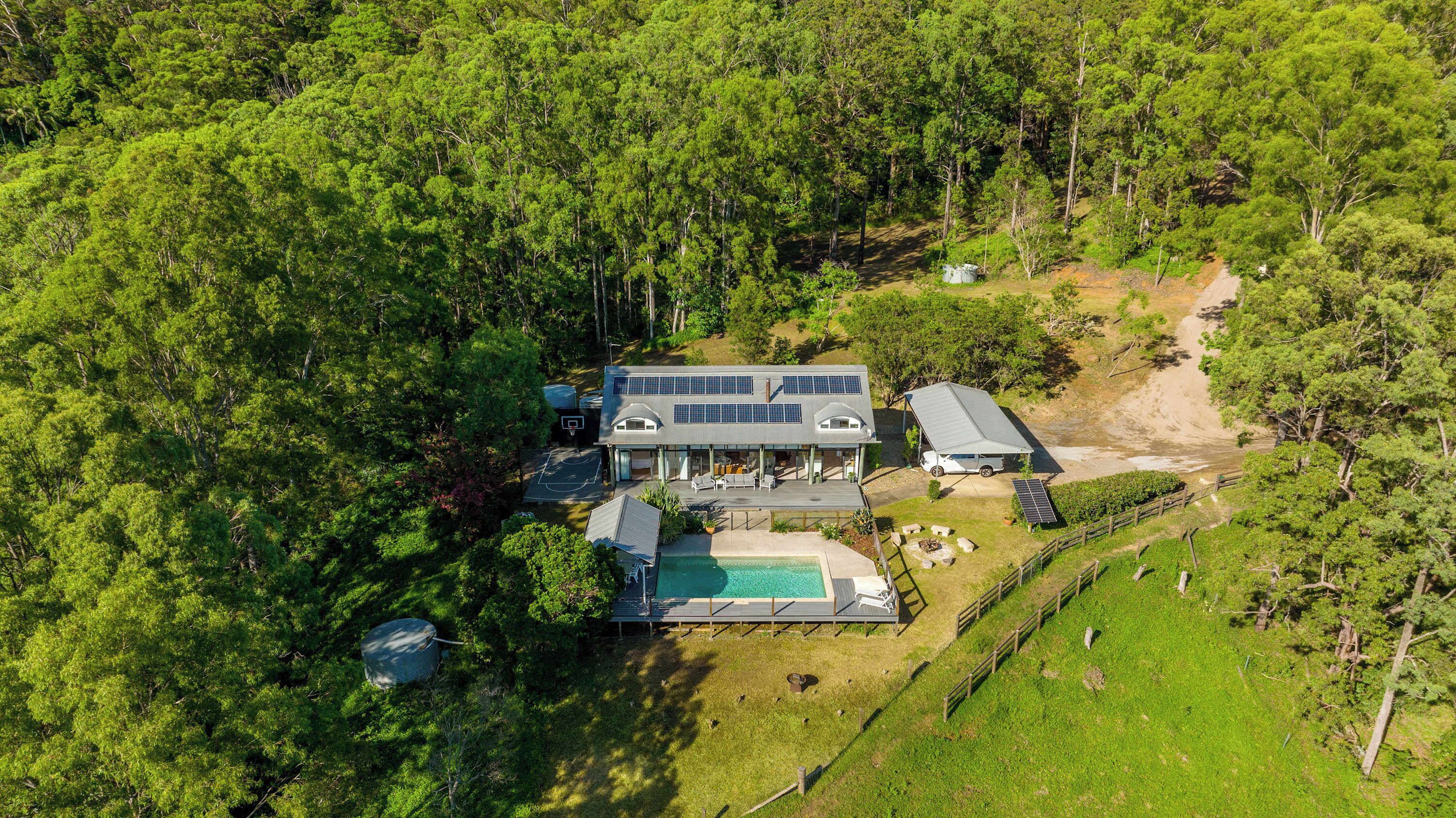 A modern house with solar panels, a pool, and a tennis court surrounded by dense forest.
