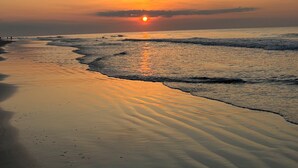 Una playa al atardecer, con el sol bajo en el horizonte, olas mansas lamiendo la orilla y un cielo despejado.