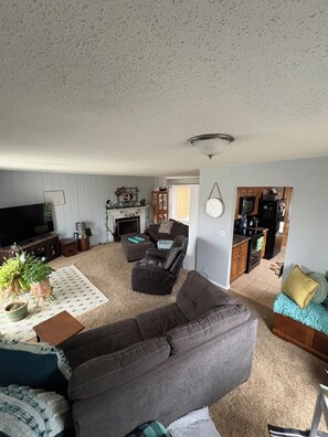 A living room with a large gray sectional sofa, a coffee table, and a TV. There is a kitchen area with cabinets and appliances visible through an open doorway. - Home available for EAA July 17-25,2026 (Oshkosh)