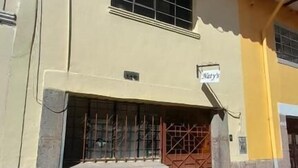 A building with a yellow facade, a barred entrance, and a sign reading "Wats". - Natys guest house (Cusco)