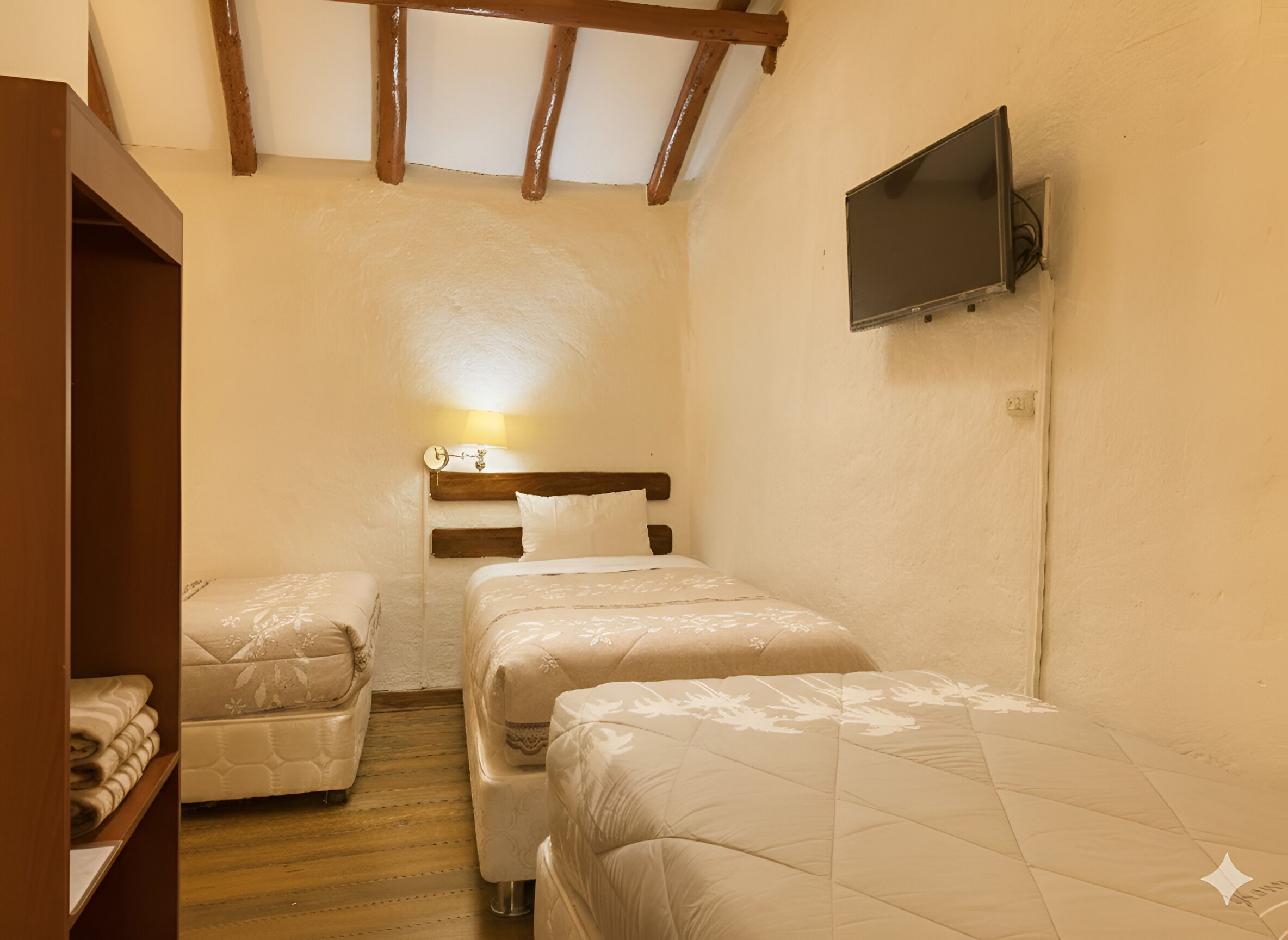 A small, cozy room with two beds, a wooden ceiling, a TV mounted on the wall, and a shelf with folded blankets.