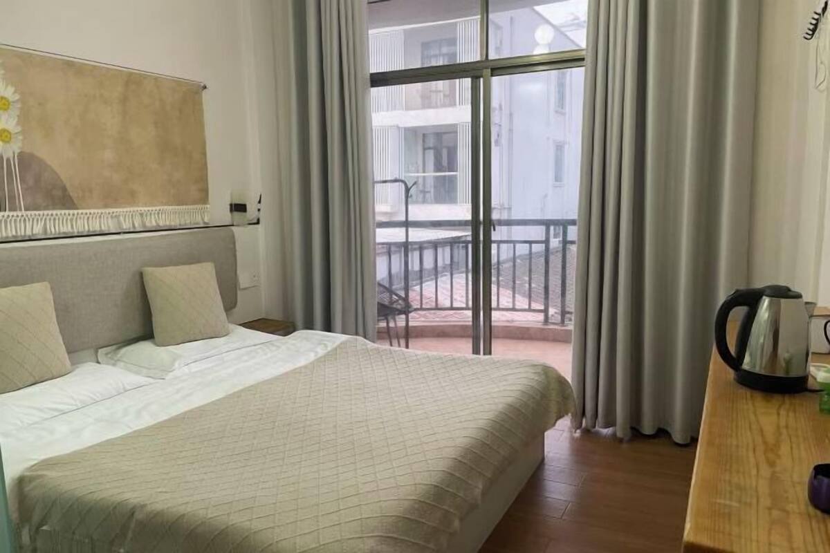 A hotel room with a bed, a view to the outside, a table with a kettle, and a painting on the wall.